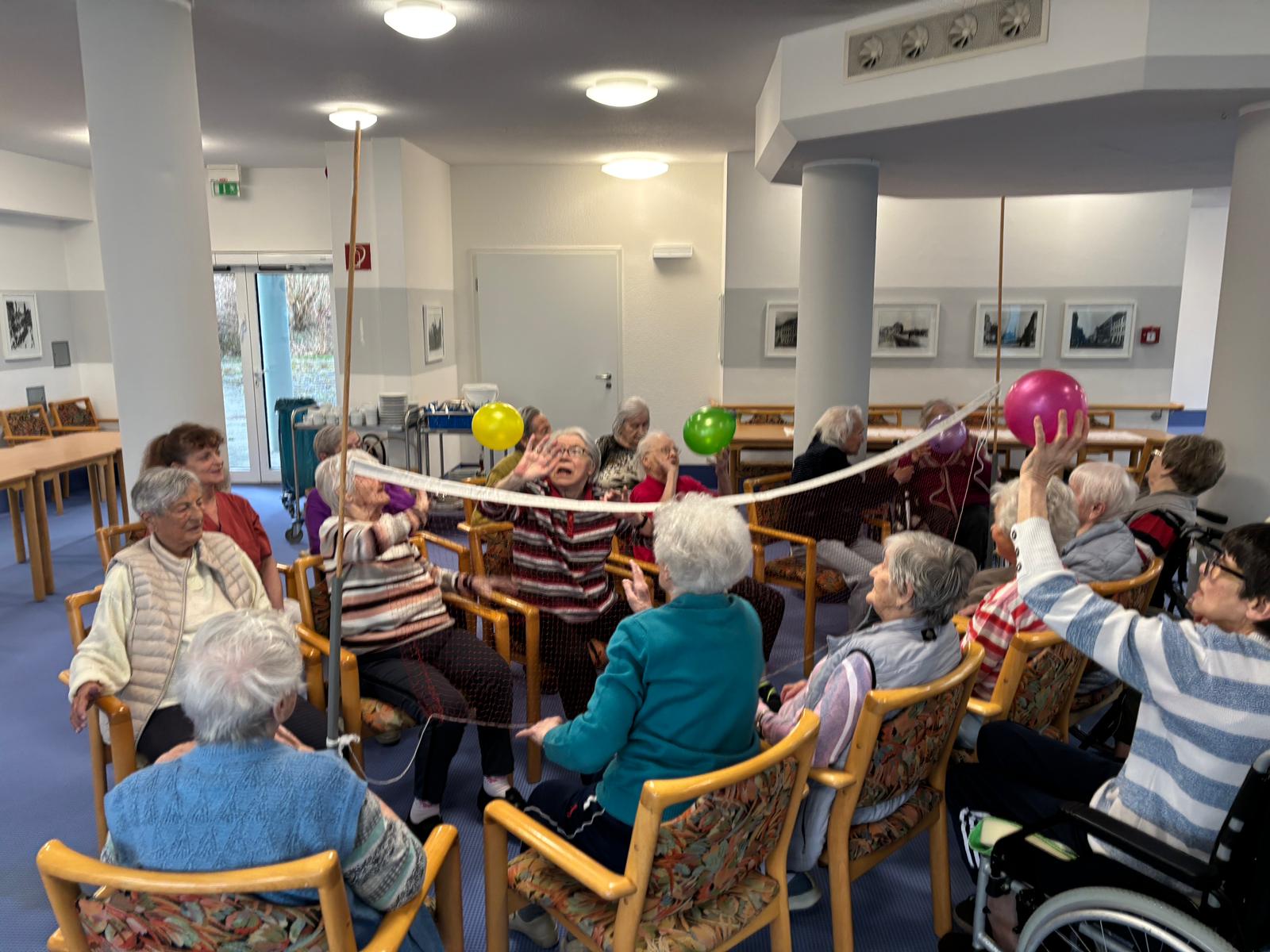 die Senior*innen sitzen sich auf Stühlen gegenüber, dazwischen ist das Volleyballnetzt, die Senior*innen werfen sich die Ballons über das Netz