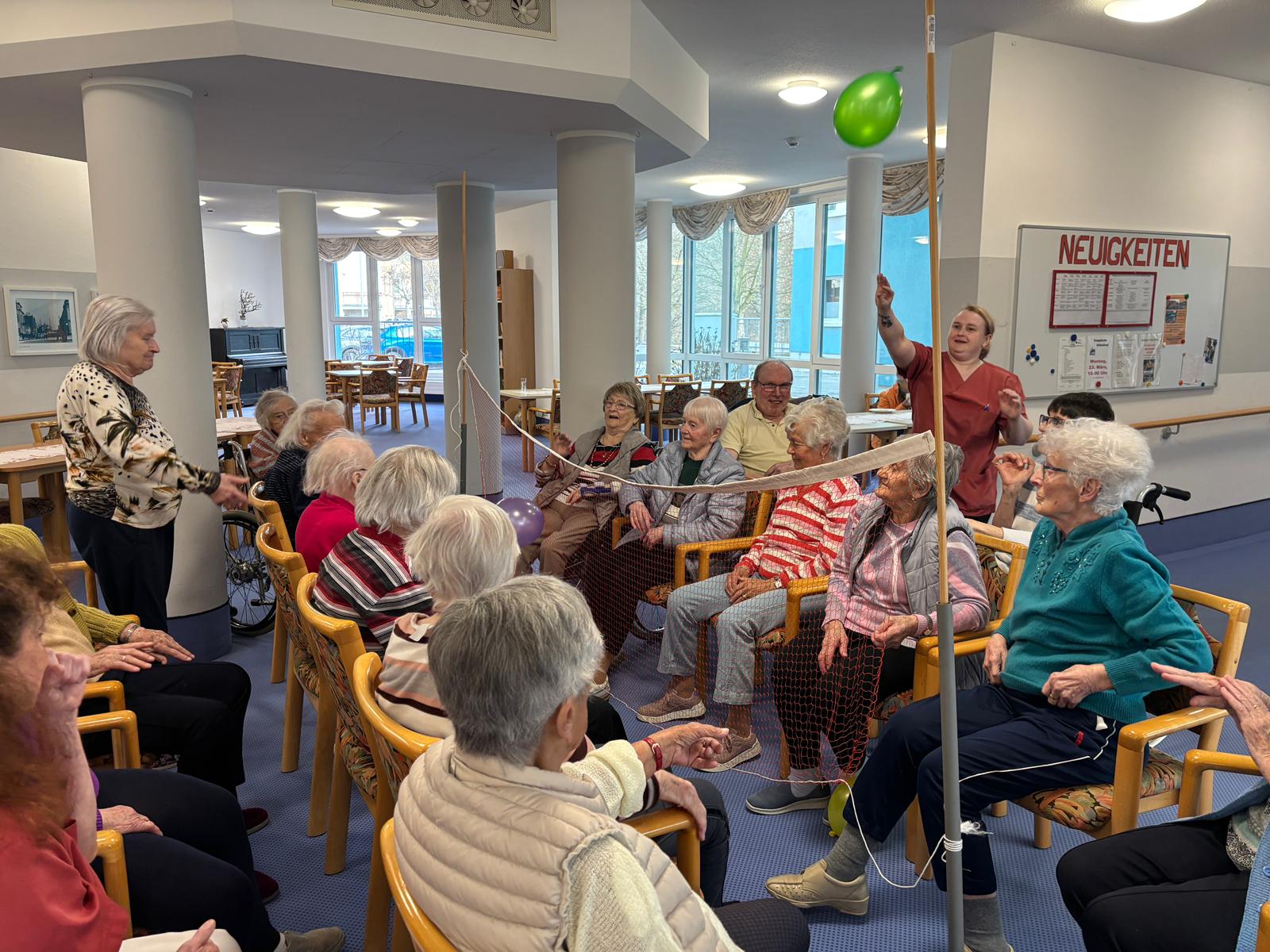 die Senior*innen sitzen sich auf Stühlen gegenüber, dazwischen ist das Volleyballnetzt, die Senior*innen werfen sich die Ballons über das Netz