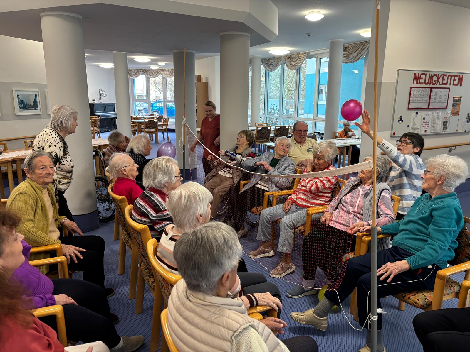 die Senior*innen sitzen sich auf Stühlen gegenüber, dazwischen ist das Volleyballnetzt, die Senior*innen werfen sich die Ballons über das Netz