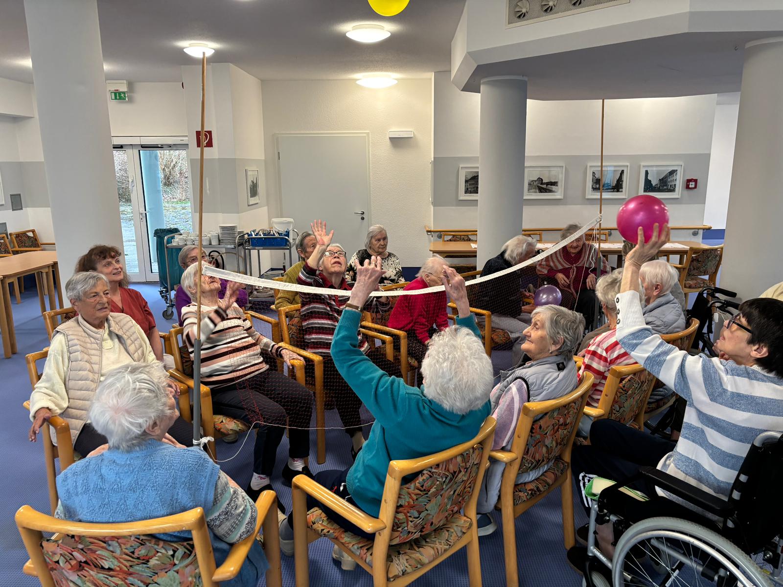 die Senior*innen sitzen sich auf Stühlen gegenüber, dazwischen ist das Volleyballnetzt, die Senior*innen werfen sich die Ballons über das Netz